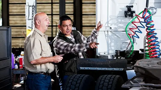 Two men engaged in a conversation, with one pointing to a specific area of a truck undergoing repairs, highlighting the importance of maintenance and repair services covered by truck financing agreements.
